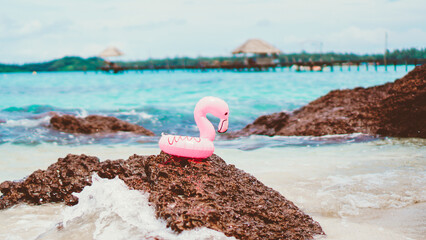 A swim ring in the shape of a pink flamingo, on the sand of a beach, with the ocean in the background. Flamingo shape, floating rubber ring by the beach.
