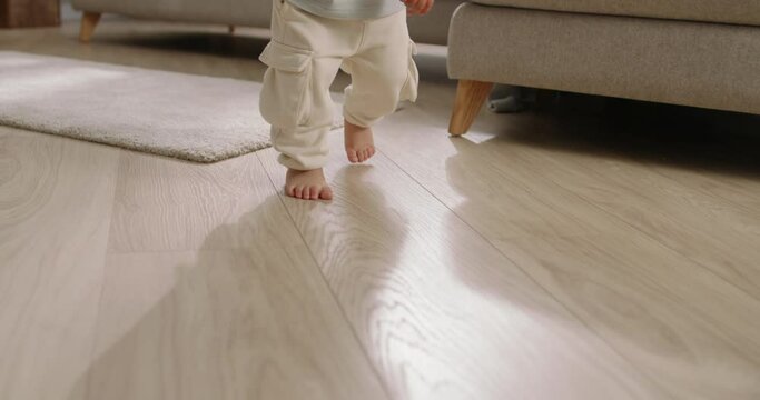 Baby first steps. Chinese, Japanese, Korean, Asian. Barefoot kid toddler walks across floor, first uneven uncertain steps in life. legs close-up. Baby's feet on floor at home. Toddler learning to walk