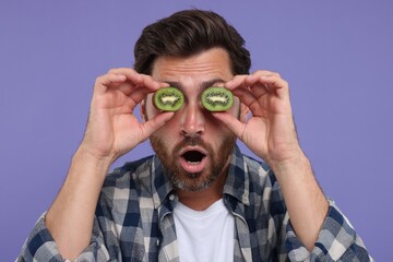 Emotional man holding halves of kiwi near his eyes on purple background