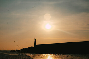 A plane fly over the white lighthouse. Lighthouse on sunset.