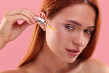 Fototapeta premium Beautiful young woman applying cosmetic serum onto her face on pink background, closeup