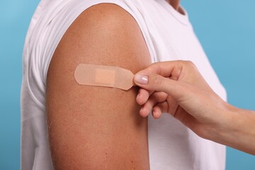 Nurse sticking plaster on man's arm after vaccination against light blue background, closeup