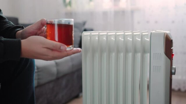 A man holds a cup of hot tea in his hands against the backdrop of an electric heater at home. The concept of heating an apartment during cold weather. Copy space for text