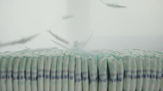 Feathers fall on a stack of baby diapers on a white background. The concept of softness and comfort, thorn-allergenic diapers made of cotton. Copy space for text. Slow motion