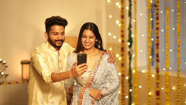 A married Indian couple happily waving and chatting on a video call during Diwali, colourful decorations. A modern young couple celebrating the occasion.