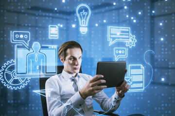 Attractive young european businessman with tablet and creative blue devices hologram sitting on blurry office interior background. Digital marketing, social network and online service concept.