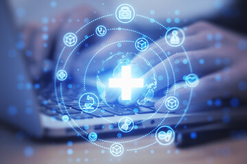Close up of male hands using laptop on desk with creative glowing blue medical hologram with cross on blurry background. Science, healthcare system and futuristic pharmacy concept. Double exposure.