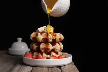 Pouring honey onto delicious Belgian waffles with strawberries and butter at wooden table against black background, space for text