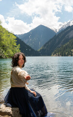 A woman in a long skirt stands on the shore of a mountain lake