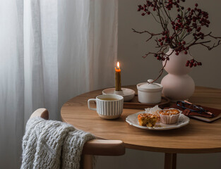 Cozy tea party - a cup of tea, muffins, cranberry branches, a bouquet in a vase, a notebook, a burning candle on a round wooden table in the living room