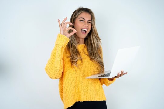 Attractive cheerful skilled Beautiful caucasian woman wearing yellow jumper using laptop showing ok-sign winkin