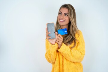 Beautiful caucasian woman wearing yellow jumper holding bank card modern device looking empty space