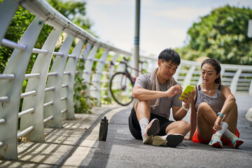 young asian couple sitting on ground looking at cellphone together while resting relaxing after exercise