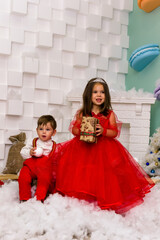 A little girl and a boy in red clothes have fun in the artificial snow by the fireplace in the studio. The girl is holding a box with gifts. Little kids in red christmas costume having fun beside a