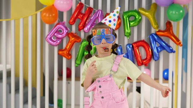 A little cheerful girl in a hat with paper glasses and a pipe on the background of a wall with balloons. Portrait of a birthday girl at a birthday party.