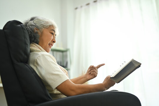 Low angle photo of beautiful senior woman sitting on armchair and reading book. Elderly lifestyle concept