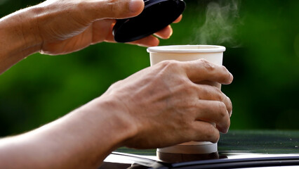 coffee takeaway in a paper cup on top of the car roof green tree background at sunrise in the...
