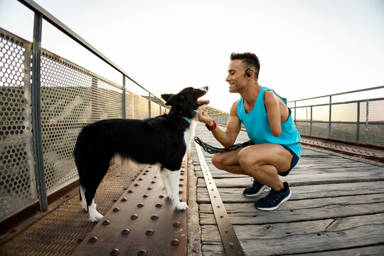 An adult athlete with an amputated arm bends down to pet his dog on an outdoor bridge. The man strokes the muzzle of a border collie. Canicross concept. Activities with animals