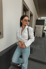 Beautiful fashion casual woman model with vintage sunglasses in a white jacket with blue high waist jeans with bag walks in the city