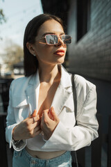 Stylish young beautiful girl with vintage sunglasses in a fashionable white blazer in the sunlight on the street. Urban pretty lady