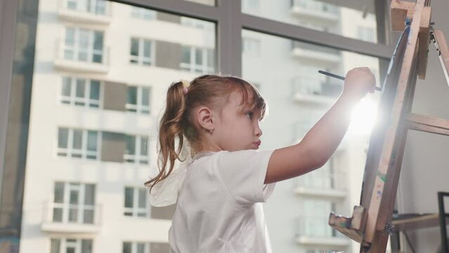 Cute girl with ponytail is painting picture with acrylic paints on canvas. Child is in room with panoramic windows. Outside another high-rise building is visible. Sun rays hitting lens create glares