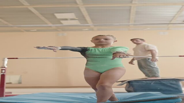 Vertical full length shot of acrobatic Caucasian little girl in green leotard performing artistic elements with gymnastics bar and jumping off on mat while training with coach indoors