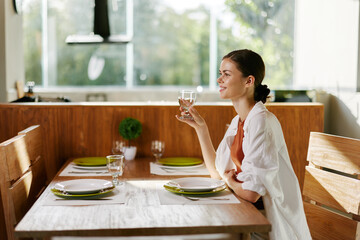 Stylish woman sitting at dining table with beautiful smile, home modern interior, sunset light, lifestyle in comfort.
