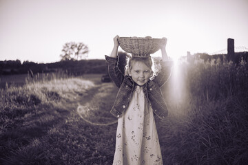 Beautiful three-year-old girl posing with a wicker basket of apples holding on her head. A little girl with a basket of apples on head in a sun-drenched meadow.