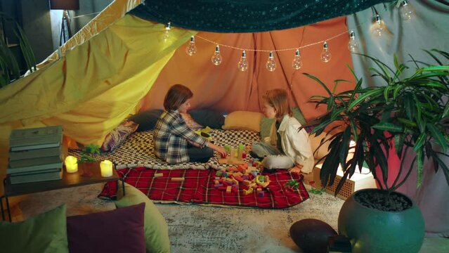 Two young boys are playing with rainbow colored building blocks in a multi colored blanket fort