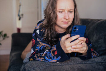Woman in sweater looking at her smartphone shocked and surprised, laying on a couch at  home