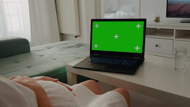 Shot of green screen laptop on coffee table and pregnant female belly sat on sofa. Pregnant tired female on the couch in front of a computer with chroma key screen..