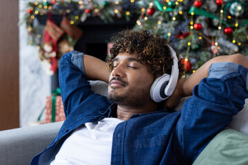 Indian young man wearing headphones, eyes closed and hands behind head sitting on sofa at home and...