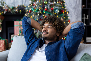 Calm and tired young Indian man in blue shirt resting on sofa at home on New Year holidays. Closing his eyes, throwing his hands behind his head, he dozes and rests. Close-up photo.