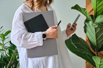 A woman holds a laptop and graphics tablet against a background of greenery.