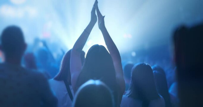 Woman clapping hands at concert, back view. Fan silhouette raises hands up in air, clapping in blue stage spotlights light. Audience applause at music band performance. Happy young girl applauding