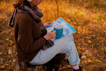 woman with a backpack drinks tea from a cup, dressed in a brown sweater, holds a map in her hands, enjoys sunny weather in the autumn forest, traveler on the precipice, hiking - obrazy, fototapety, plakaty