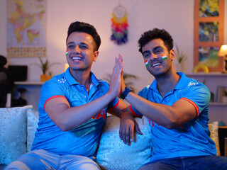 Random shot of two men giving high five while watching a one-day match at home - wearing a cricket jersey . Indian Friends enjoying the cricket match at home - sitting on a couch cricket lovers c...
