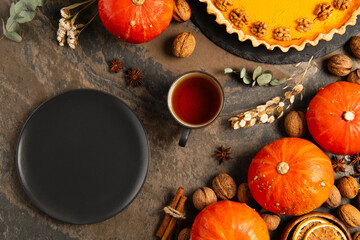 black ceramic plate near orange pumpkins, tea and thanksgiving pumpkin pie on rustic stone tabletop