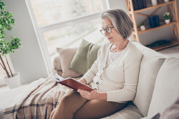 Obraz premium Photo of cheerful charming elderly lady wear white cardigan sitting sofa reading interesting book indoors apartment room