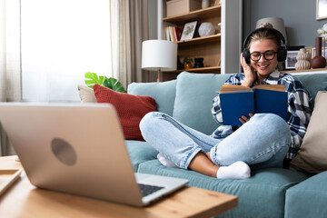 Young beautiful hipster woman sitting on sofa at home listening music on wireless headset and reading a book. Freelance female enjoy free time after finishing work on laptop, read new novel bestseller - obrazy, fototapety, plakaty
