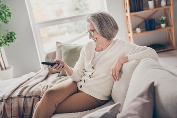 Photo of pretty cheerful elderly lady wear white cardigan sitting sofa watching television indoors...