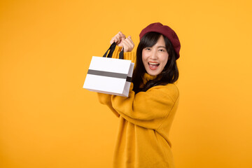 Young Asian woman in her 30s, with a shopping paper bag, displaying elegance in a yellow sweater and red beret during her shopping spree against a yellow background.