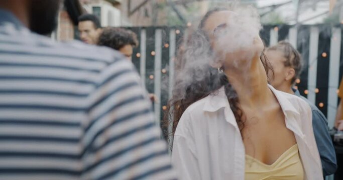 Slow motion of happy young woman smoking electronic cigarette and dancing at street party with friends. Vaping device and modern lifestyle concept.