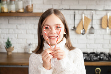 Close-up of child hiding behind two candy canes in shape of heart in kitchen setting. Kid smiling and looking at camera. Blurred background with white brick wall and kitchen utensils hanging on it