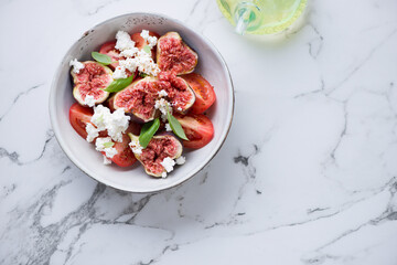 Bowl of fig fruit, tomato and goat cheese salad on a white marble background, horizontal shot with space, flat lay