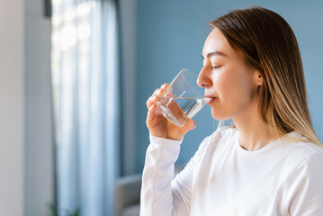 Woman drinks water from a glass in the living room. Concept of a healthy lifestyle and women's wellness.