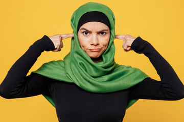 Young arabian asian muslim woman wear green hijab abaya black clothes cover ears with hand finger do not want listen scream isolated on plain yellow background People uae middle eastern islam concept
