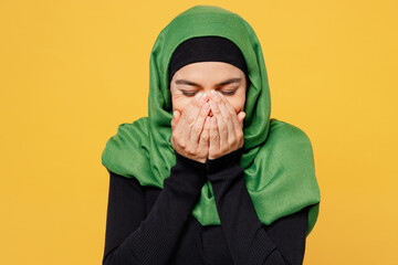 Young arabian asian muslim woman wearing green hijab abaya black clothes cover mouth face with arms hands palms isolated on plain yellow background. People uae middle eastern islam religious concept.