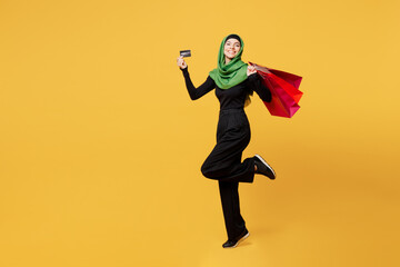 Fototapeta na wymiar Full body young amazed asian muslim woman wears green hijab abaya clothes hold shopping package bags credit bank card isolated on plain yellow background. Black Friday sale buy day, uae islam concept.