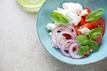 Salad with red tomatoes, feta cheese, red onion and fresh green basil in a turquoise bowl, horizontal shot on a beige stone surface